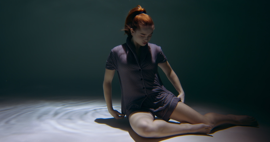 Young beautiful peaceful redhead woman wearing night pyjamas sits down deep under water on dark pool floor slow motion.