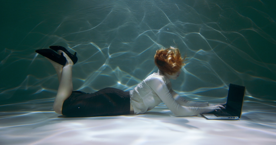 TAKING A BREAK. Young redhead business woman using laptop deep under water, swimming up to rest from work slow motion.