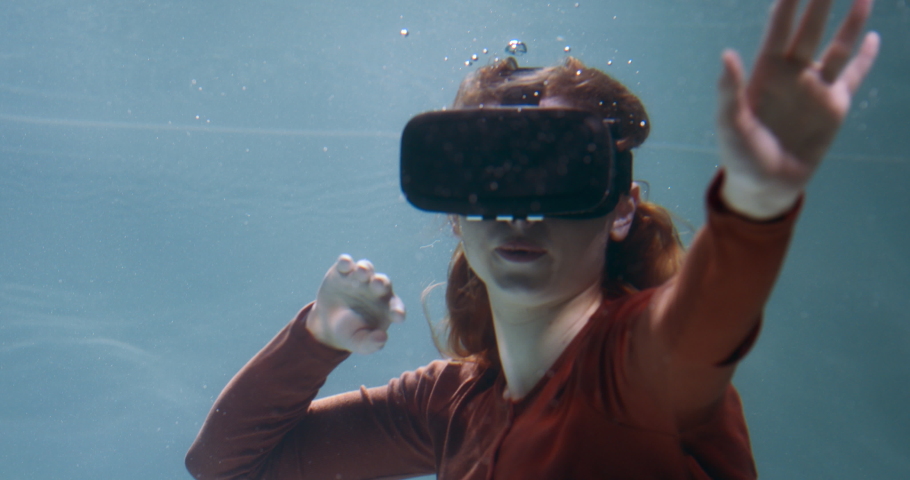 Immersed in virtual reality. Young beautiful smiling redhead woman using VR headset floating under water slow motion.