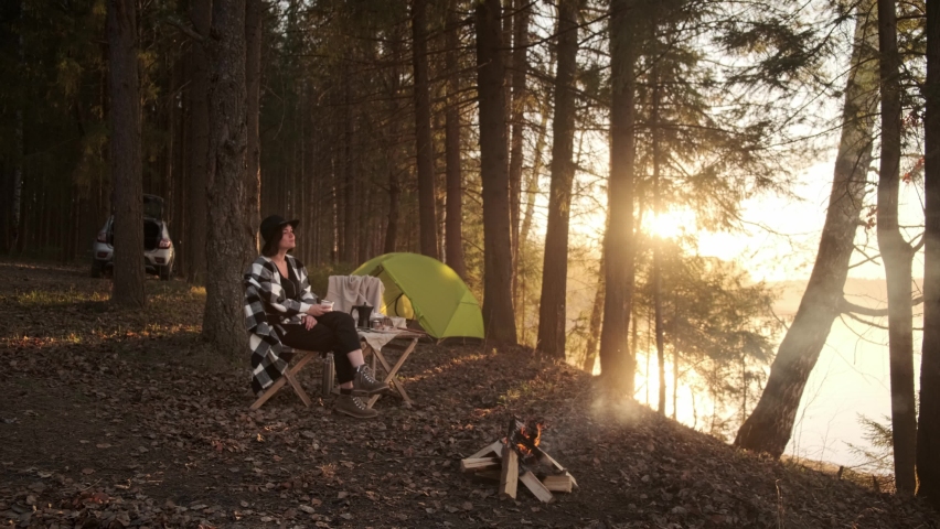 A young girl on a picnic with a tent fries marshmallows over the fire and drinks coffee.