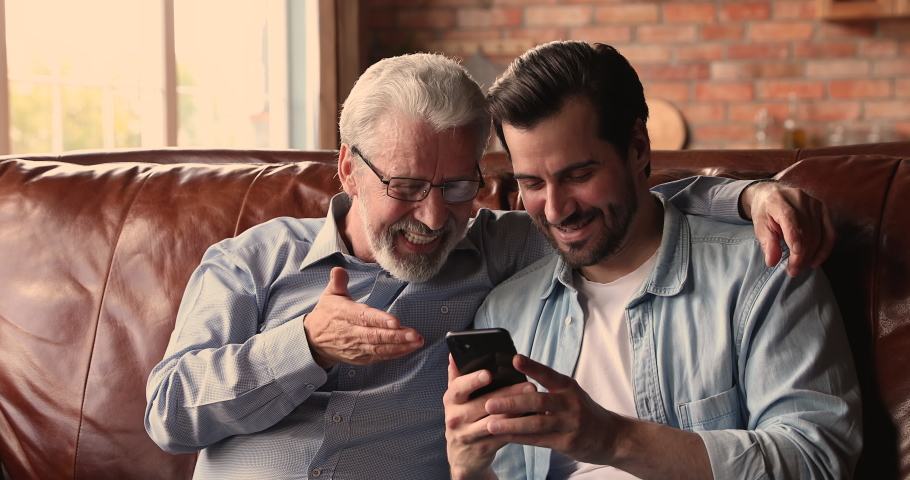 Millennial man sit on sofa hold smartphone showing to older dad new mobile application, explain usage of most useful medical apps for seniors, discussing media news. Modern tech and elderlies concept Royalty-Free Stock Footage #1072477994