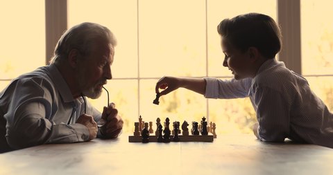 Older grandpa and little 7s grandkid play chess sit at table at home. Small boy aged grandfather spend time together talking making moves enjoy board game. Leisure activity, hobby, brain work concept