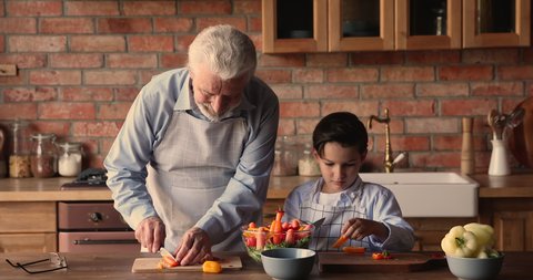 Elderly grandpa and little grandson wear aprons cooking together in kitchen, cutting on wooden boards peppers talking spend time at home. Cookery, hobby, multigenerational family prepare food concept