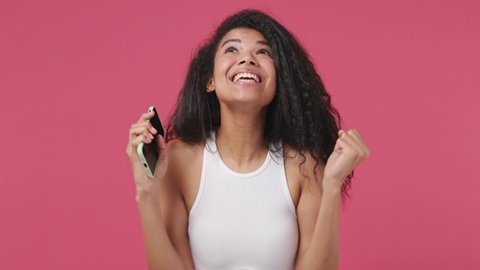 Young excited african woman wear white tank top shirt hold talk use mobile cell phone just found out great big win news doing winner gesture say yes isolated on dark pink color wall background studio