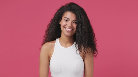Young african woman curly hair 20s years old wears white tank top shirt posing look camera charming smile isolated on dark pink color wall background studio. People sincere emotions lifestyle concept