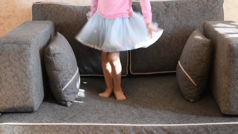 3-5s little girl in blue tulle tutu skirt with barefoot legs jump on grey couch with pillows. Kid having fun at home. Happy childhood