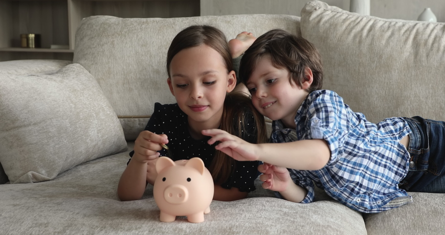 Cute smiling little brother and sister lying on couch and take turns dropping coins into piggy bank, save pocket cash for fun, dreams and future education. Keep money, thinking about tomorrow concept Royalty-Free Stock Footage #1072483667