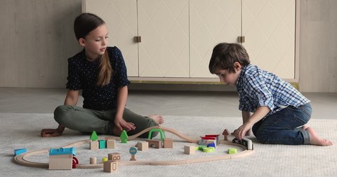 Siblings play seated on warm floor in living room. Little boy and girl enjoy modern wooden plaything, riding train on toy railroad set. Hobby and leisure at home, interesting childhood games concept