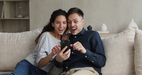 Ecstatic millennial Hispanic couple sit on couch check mailbox use mobile phone read great news celebrate success, giving high five gesture enjoy moment of victory showing sincere emotion of happiness