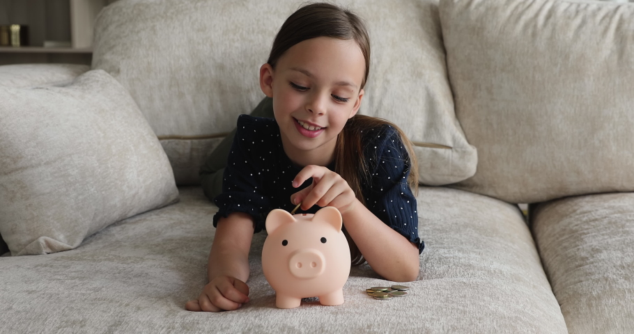 Beautiful 7s girl lying on sofa counts and dropping coins into piggy bank. Save money, pocket cash for future needs, education, purchases, learn kid be thrifty, economy skills from childhood concept Royalty-Free Stock Footage #1072486994