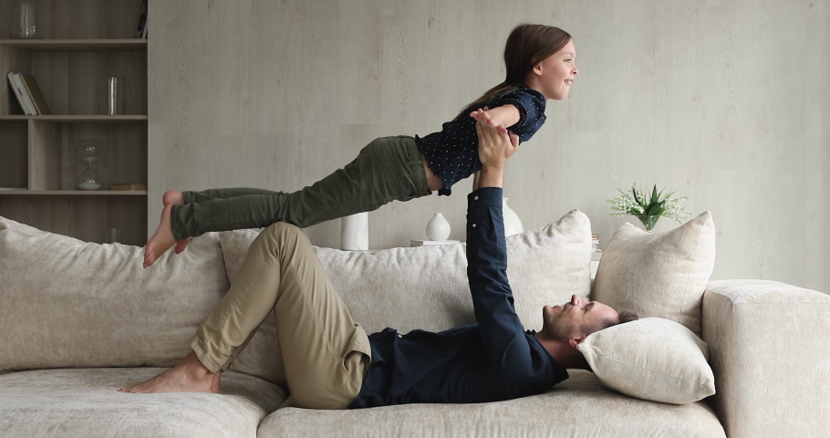 In living room strong dad lying on sofa holding lifting little daughter on outstretched hands, child spread arms like plane wings imagines like fly in air. Sportive family, yoga practice, fun concept Royalty-Free Stock Footage #1072487000