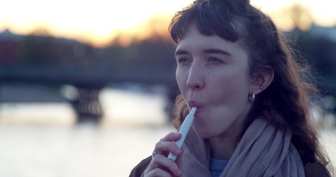 portrait of a girl smoking e-cigarettes in the street against the water and the sun leaving in the evening