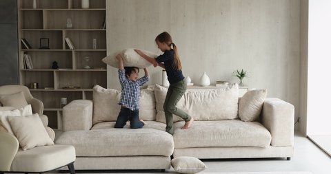 Cheery siblings play tag game in living room, little sister catching running chasing preschool brother active kids jump on sofa play with cushion having fun together at home. Weekend playtime concept