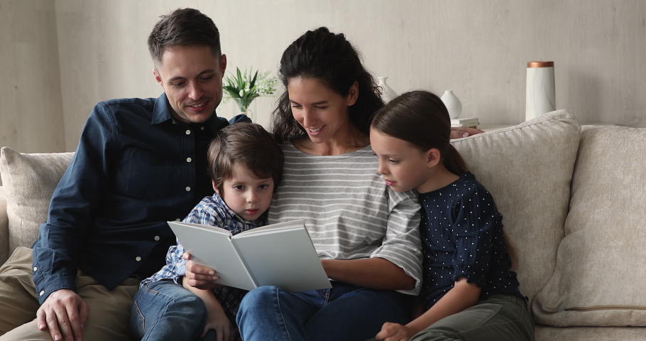 Young loving parents reading fairy tale book to little beautiful son and daughter, family sitting on comfortable couch in living room enjoy interesting story. Children development, upbringing concept Royalty-Free Stock Footage #1072493204