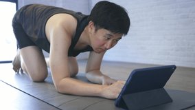 Young Asian man exercising at home while watching a video tutorial on tablet. Male fitness guy training indoors online