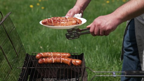 Man on picnic prepares juicy sausages in barbecue. Chef putting ready roasted hot dog sausages into a plate in the garden.