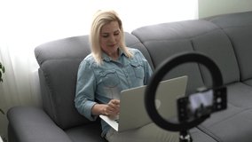 Smiling woman recording her video blog. Adult woman speaks in front of a video camera for her blog channel. Woman blogger videotapes her vlog at home. Vlogger makes online streaming using smartphone.