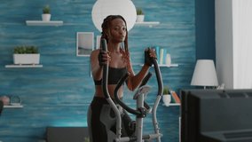 Fit athletic black woman running on elliptical bike watching wellness routine fitness video on tv during gym morning workout in living room. Athletic black woman training body muscle