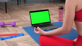 Green screen mock up chroma key monitor laptop: Calm sportive woman in sportswear practices yoga online video call webcam computer at home. Sports girl sits meditating in lotus position on fitness mat