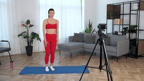 Sport muscular beautiful girl trains teaches student records online course lesson webinar on camera. Fit woman fitness trainer blogger in sportswear meditates while standing on yoga mat at modern home