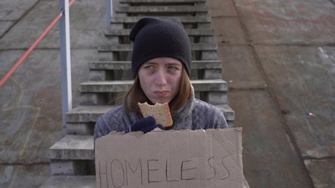homeless young woman eats bread sitting on stairs wrapped in a blanket. No war homeless girl with dirty face and gloves needs help. Social problems, Homeless people, refugee problems, escape from war