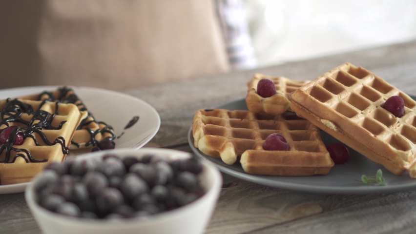 Two plates of waffles. A girl pours chocolate sauce over Belgian waffles. Sweet Belgian waffles with berries. White and gray dishes on a wooden textured kitchen table. A girl in a beige apron prepares | Shutterstock HD Video #1072525274
