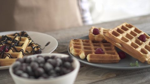 Two plates of waffles. A girl pours chocolate sauce over Belgian waffles. Sweet Belgian waffles with berries. White and gray dishes on a wooden textured kitchen table. A girl in a beige apron prepares