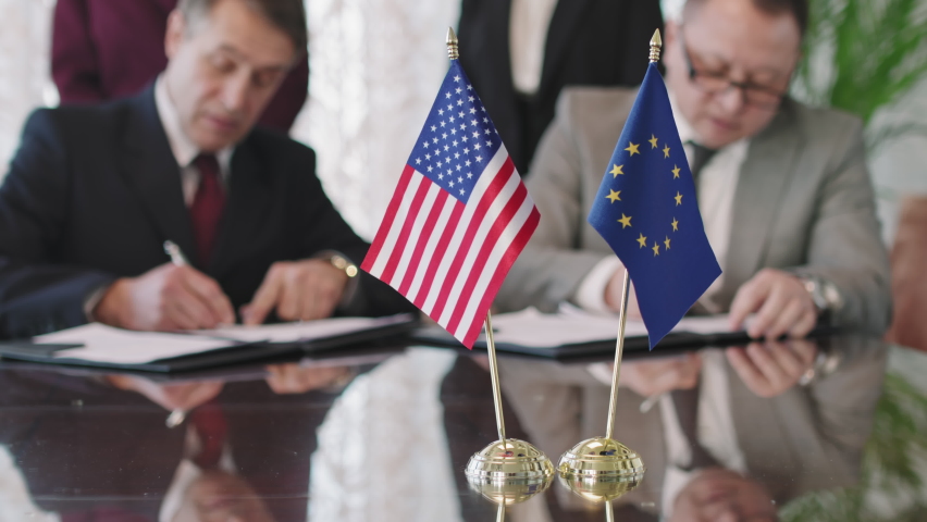 Close-up of US and EU flags on negotiation table in selective focus while political representatives of these countries signing cooperation agreement in blurred background Royalty-Free Stock Footage #1072527086