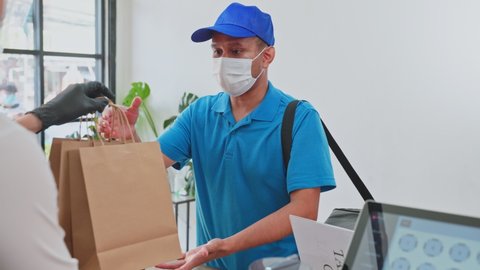 Asian Food deliverly man wearing protective mask due to Covid-19 pandemic, receive beverage order from restaurant worker at coffee shop. Waiter give takeaway bag to postman. Distance shopping concept.
