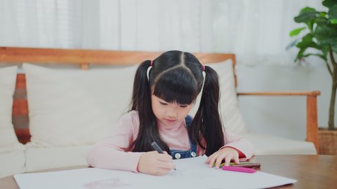 Asian small little kid daughter having fun spending free leisure time painting on paper. Young preschool girl artist playing alone drawing and coloring picture with color pencil in living room at home