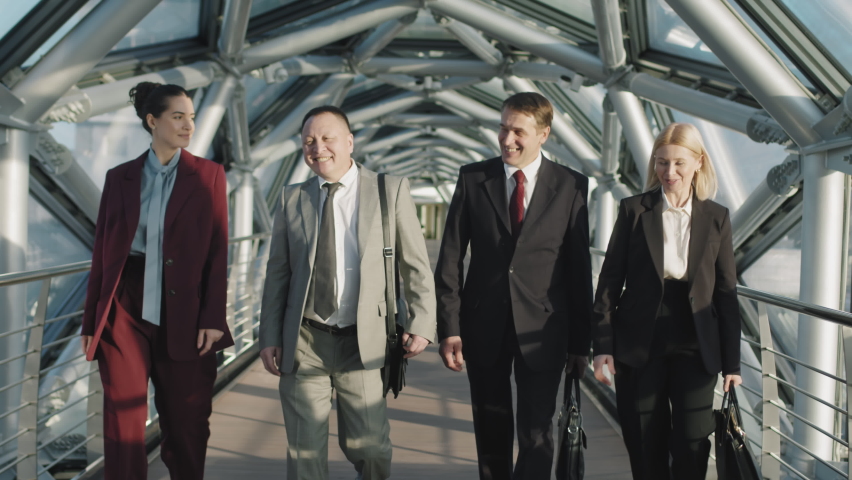 Front-view medium slowmo of successful multi-ethnic business team smiling while walking towards camera along indoor glass bridge of high-class office building