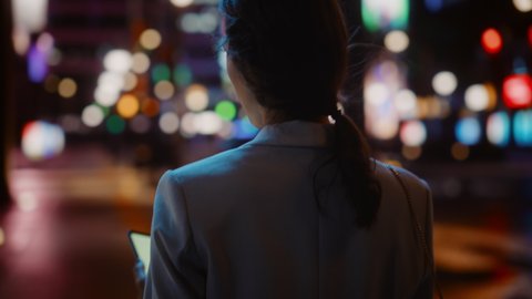 Following Shot of Beautiful Woman Using Smartphone Walking in a Modern City Street with Neon Lights at Night. Confident Female Walking Through Urban Area using Mobile Phone. Medium Tracking Cinematic