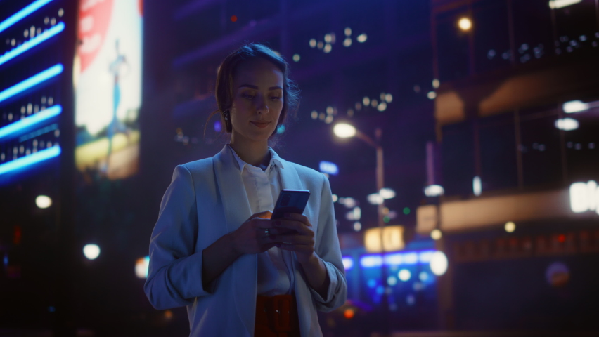 Beautiful Woman Standing, Using Smartphone on a City Street with Neon Bokeh Lights Shining at Night. Confident Smiling Beautiful Female using Mobile Phone. Medium Tracking Cinematic Flare Shot Royalty-Free Stock Footage #1072530284