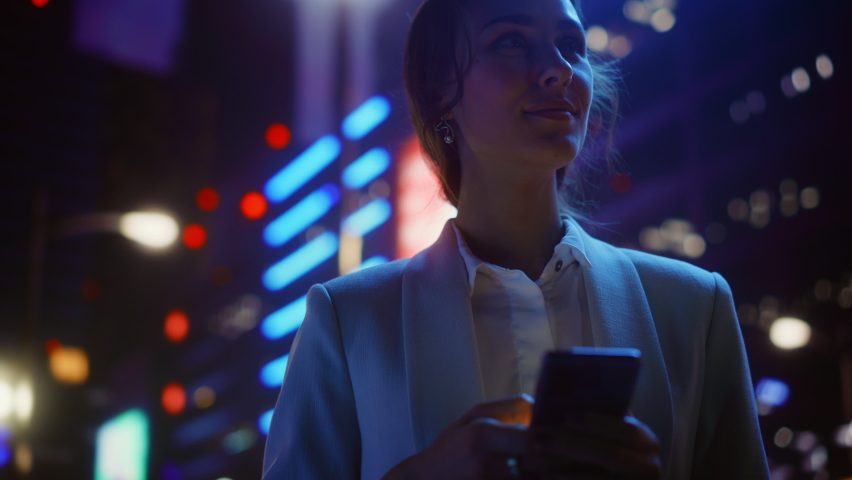Beautiful Woman Standing, Using Smartphone on a City Street with Neon Bokeh Lights Shining at Night. Confident Smiling Beautiful Female using Mobile Phone. Medium Tracking Cinematic Flare Shot