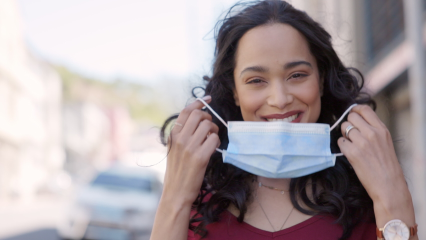 Happy Hispanic girl walking outdoor while taking off her protective face mask and looking at camera after immunization from Covid19. Young Mexican woman removing mask after receiving her vaccine shot. | Shutterstock HD Video #1072531853