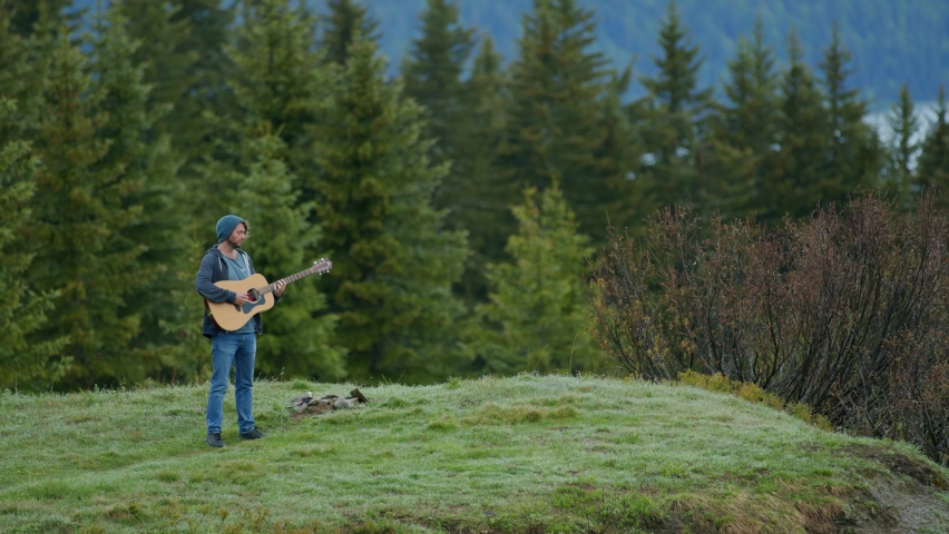 A man is playing the guitar at the top of a mountain surrounded by a forest