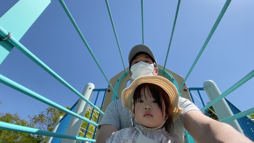 Asian family is playing on the slide. 3 years old daughter and father in medical white mask are playing on the slide together. Child looks a little uneasy. Spring season. Leisure, Activity, Outdoor