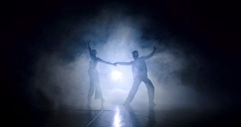 The silhouettes of a couple dancing rumba in the smoke. A spotlight is on behind them. The man holds the woman by the hand and twists towards him, then supports her by the shoulders and makes the sway