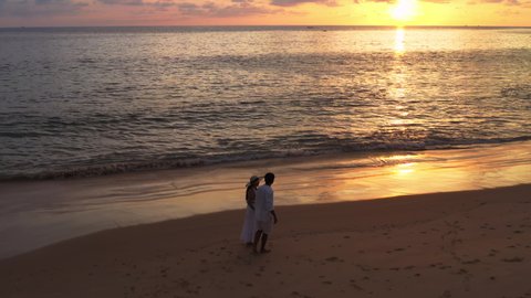4K Aerial drone top view shot of happy couple walking together on the beach with sunset over sea. Tourist couple walking on tropical beach. Romantic time on freedom paradise beach vacation holiday.