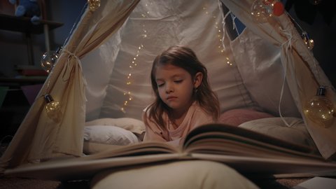 Adorable girl in cute decorative tent turning pages at home in evening. Little kid lying on floor while reading interesting book. Concept of leisure and careless childhood