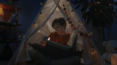 Young smart boy with blond hair in decorative tent reading interesting book at home in evening. Teenager sitting on floor while spending free time. Concept of leisure