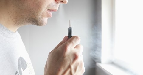 Close-up man with electronic cigarette near the window. Male smoking tobacco stick heating and exhailing smoke.