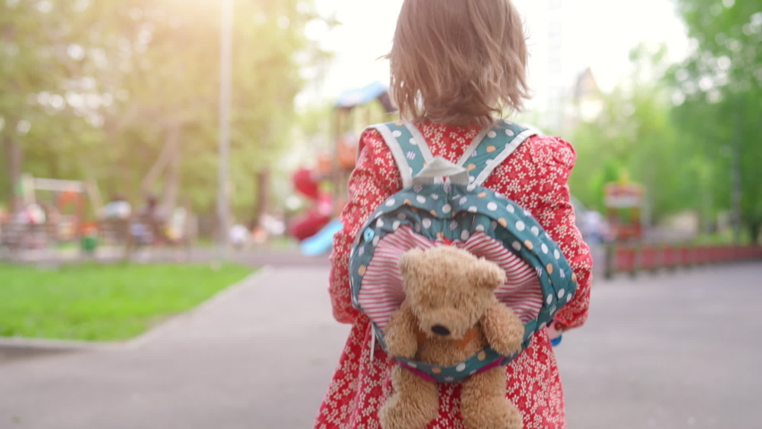 Adorable little toddler girl walking in city park with toy in backpack on her back. Childcare and active child concept