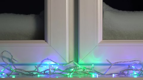 Closeup 4k stock video footage of bright colourful vivid garland holiday lights shining and glowing in darkness laying on white windowsill of white snow covered window. Christmas season