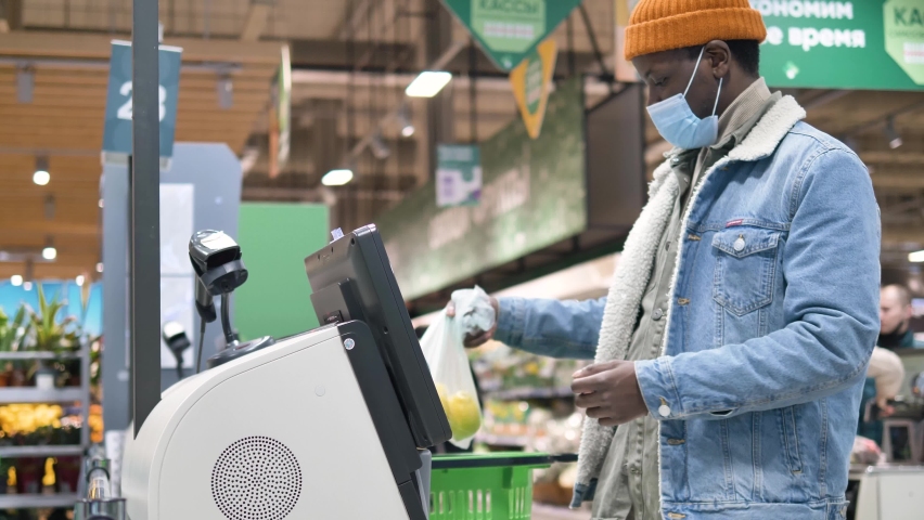 African-American man in a medical mask scans the barcode of fresh fruit at the self-service checkout in a grocery supermarket, pays with a contactless payment at the checkout of the store. | Shutterstock HD Video #1072570229