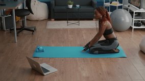 Flexible woman with black skin wearing sportswear practicing yoga sport exercises on fitness map in living room watching online pilates video using laptop. Healthy person doing morning gymnastics