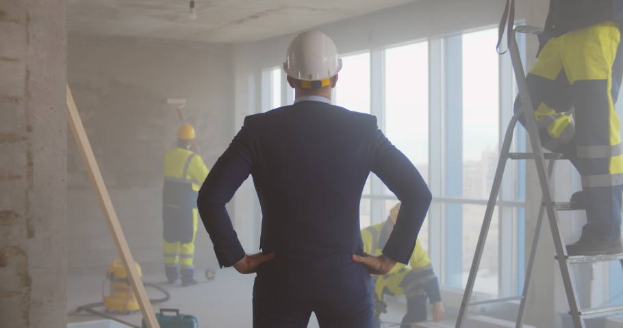 Back view of contractor in suit and hardhat supervising team of builders renovating office. Architect or owner standing in redecorating apartment watching construction workers Royalty-Free Stock Footage #1072572275
