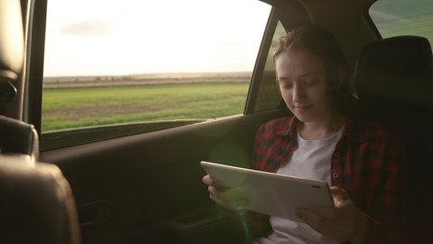 Young girl passenger travels by car with a digital tablet, sitting in back seat of a car, an open car window. Student is studying, working remotely. Free beautiful woman enjoying car ride and resting.