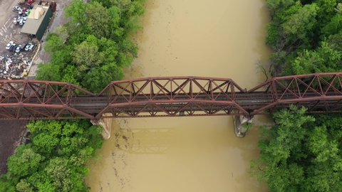 Aerial shot of a rusted brown railroad bridge over a muddy brown river surrounded by lush green tress in the summertime. Bridge crossing the Licking River from Newport into Covington.