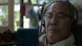 Asian senior retired mature man in eyeglasses wearing headphone enjoy listening music or watching social video from mobile smartphone in the room. Relax elderly healthy lifestyle. 